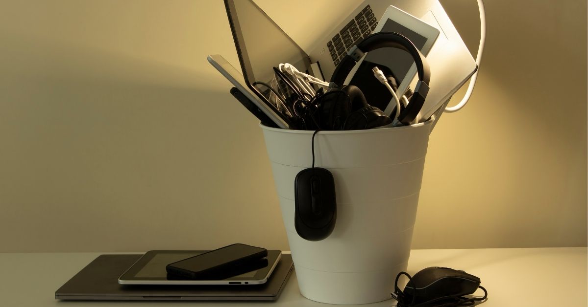 A staged photo of a garbage can filled with electronics, including a laptop computer, small tablet, large headphone, and a computer mouse dangling out.