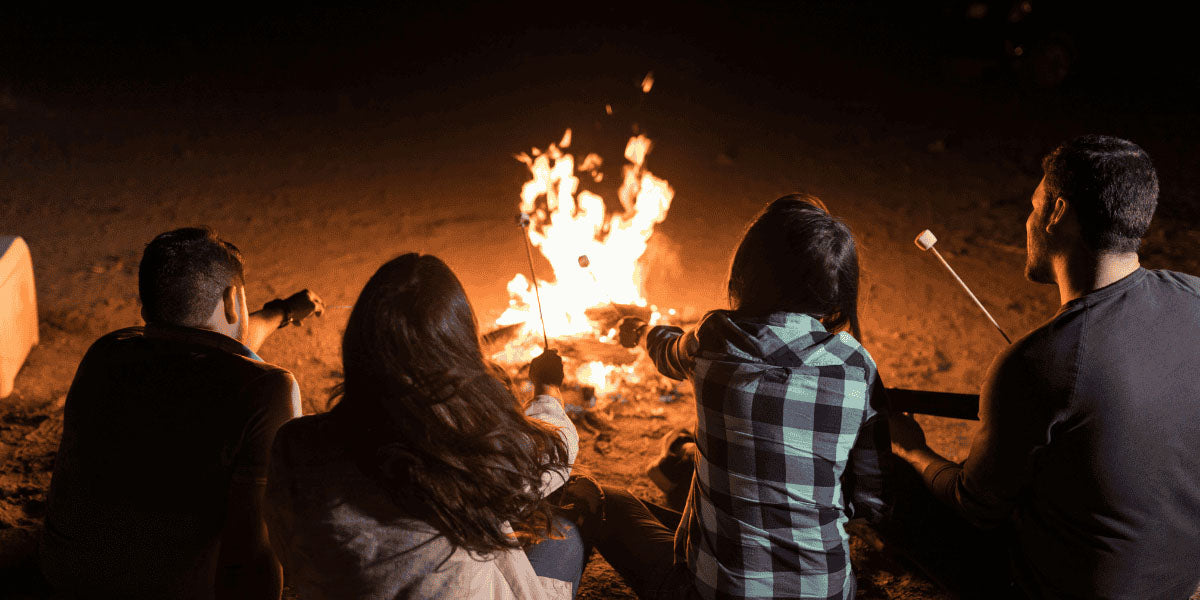 A photo showing a bonfire on a beach, with logs burning, and five different people roasting marshmallows on sticks. Everyone is sitting in the sand, so only feet, hands, and torsos are visible.