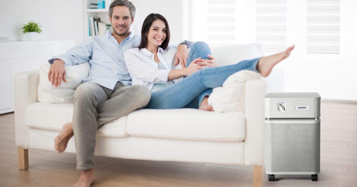 A man and woman sit together on a white couch, cozying up to each other. The room is brightly lit with sunlight. The decor is white.There is a white Austin Air purifier is in the foreground, on the right.