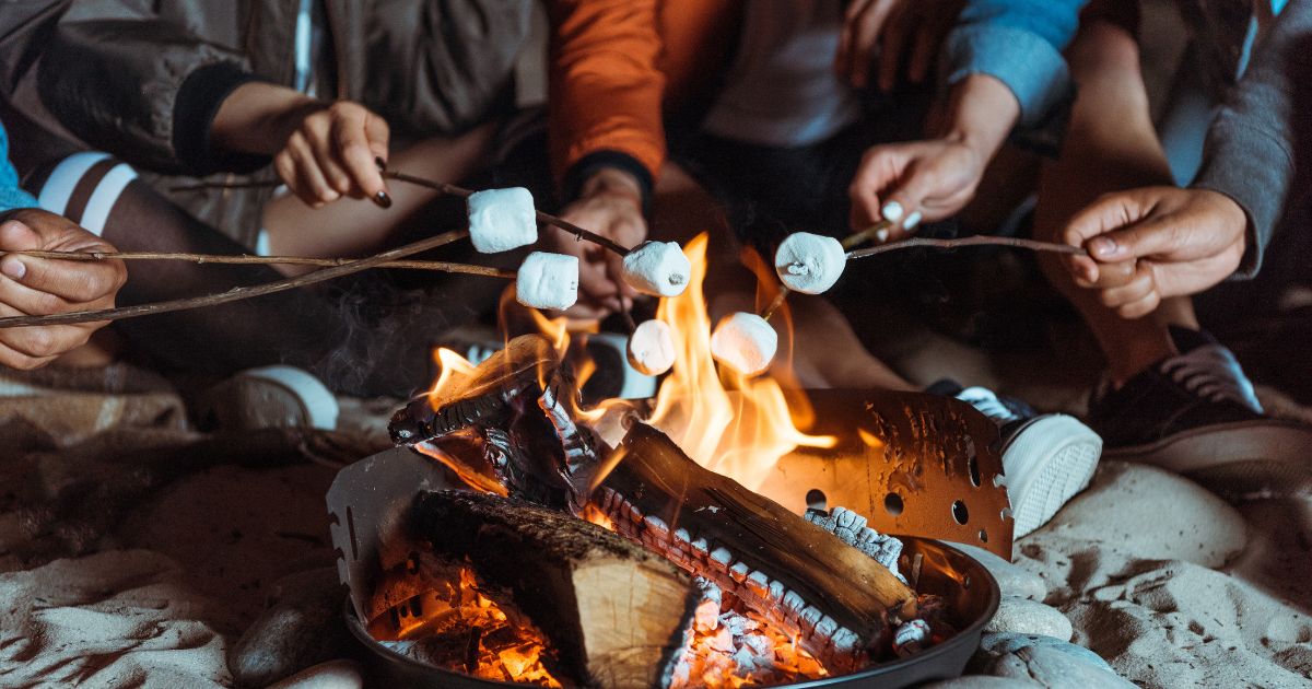 A photo showing a bonfire on a beach, with logs burning, and five different people roasting marshmallows on sticks. Everyone is sitting in the sand, so only feet, hands, and some torsos are visible.