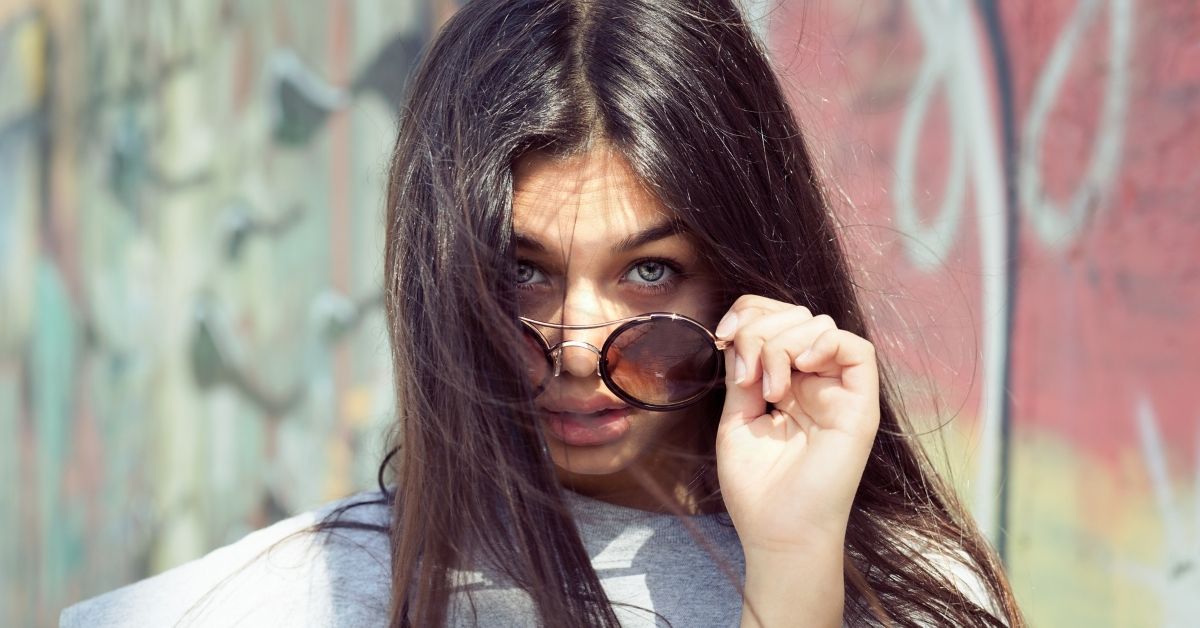 A photo of a young woman in an urban environment, graffiti is visible on the walls behind her. She has her hand on her sunglasses, lowering them and looking forward intensely.