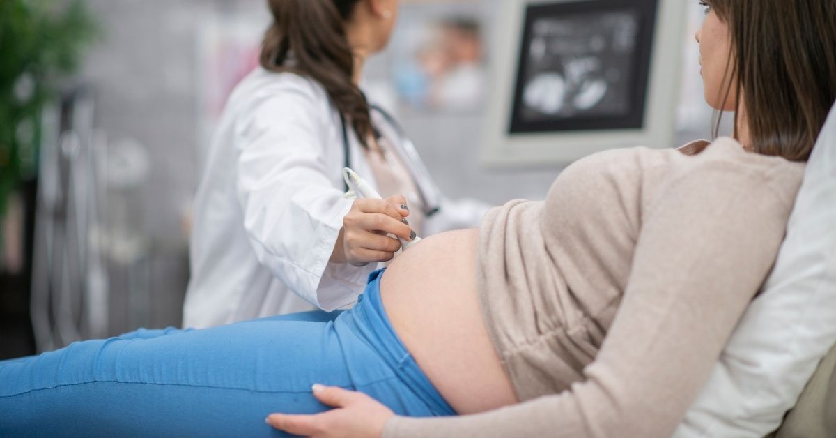 A photo, in sideview, showing a pregnant woman getting a sonogram. The technician conducting it is in the background and the screen with the baby is visible too.