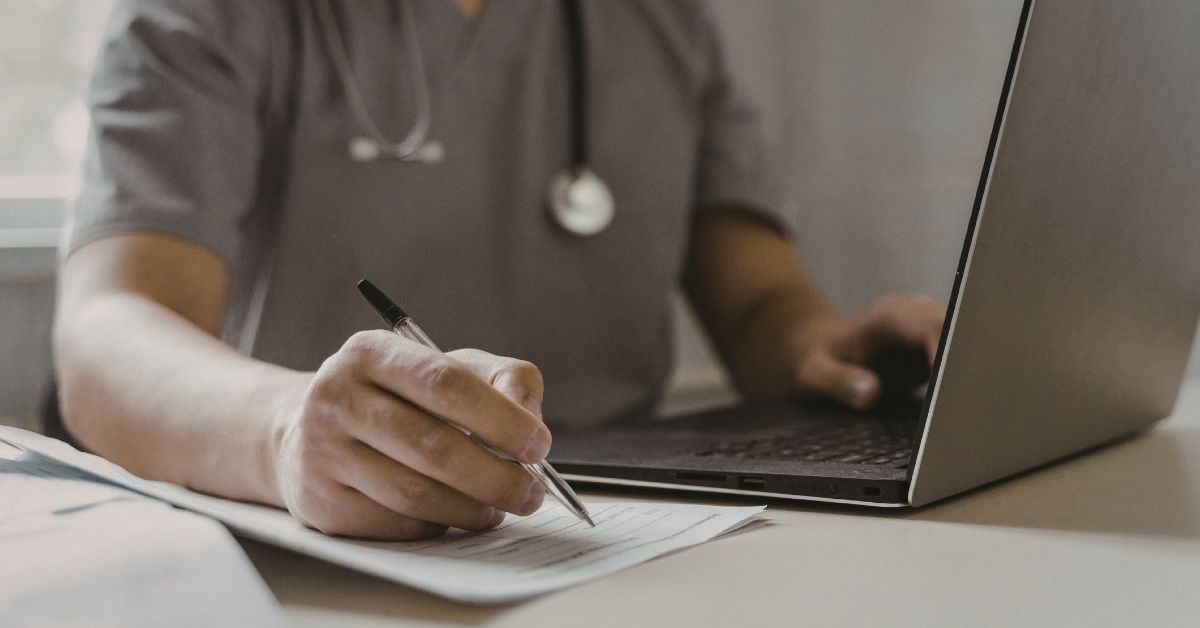 A photo showing the torso and arms of a man in gray medical scrubs who is sitting at a laptop and taking notes by hand to promote the Austin Air Mold Solutions Summit, which will be available online March 31, 2026.