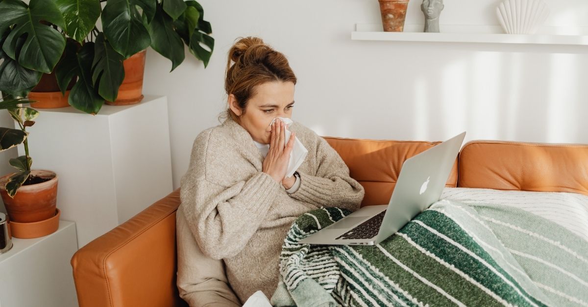 A photo of a woman sitting on a brown leather couch. She is under a blanket and blowing her nose while looking into a laptop.