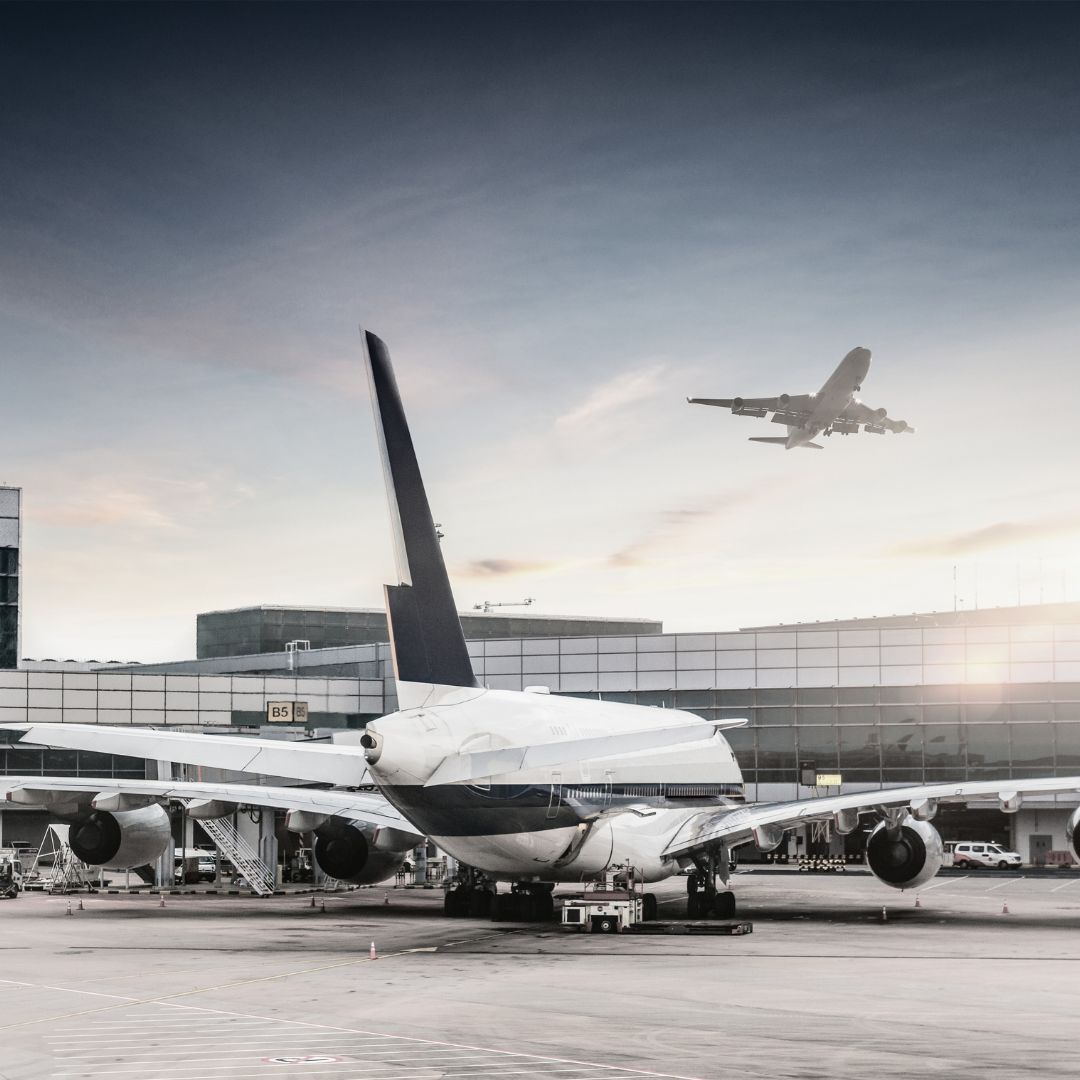 Airplane on the tarmac at an airport with another airplane in the sky.