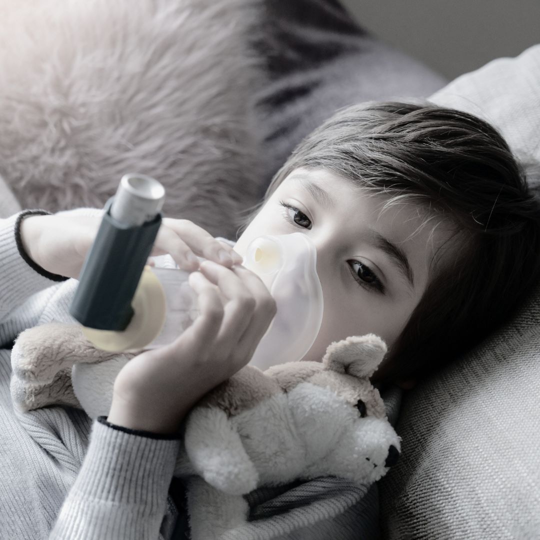 Child holding a teddy bear and an inhaler, lying on a couch.
