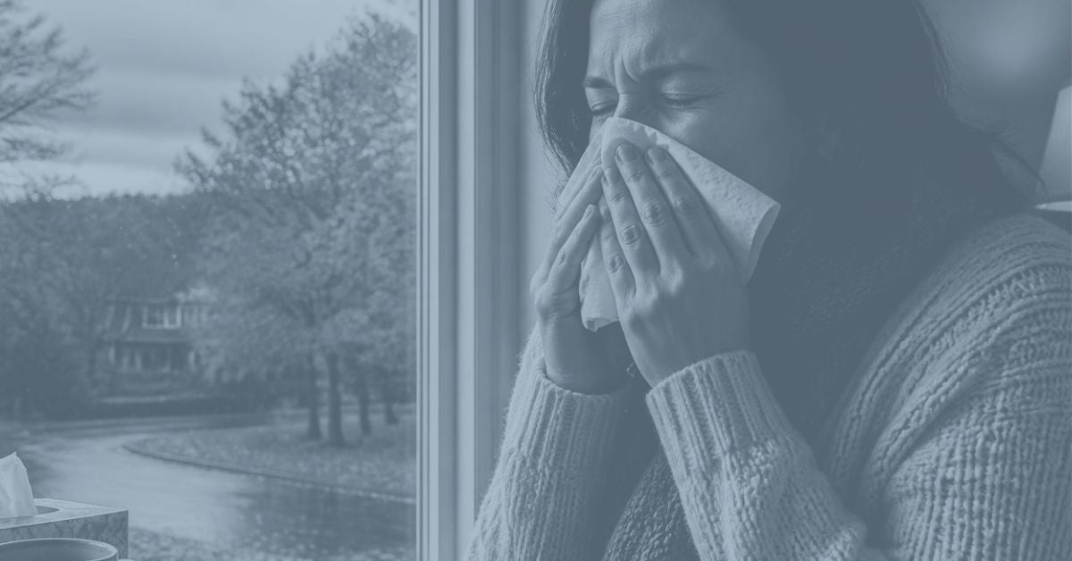 A woman with a cold or flu symptoms, holding a tissue to her nose while sitting beside a window on a cold winter day. The image has a cool, muted blue-grey tone.