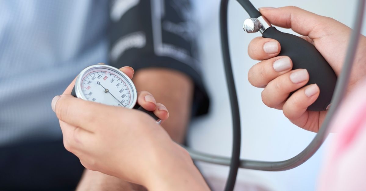 A close up photo of someone taking another person’s blood pressure. The blood pressure pump and gauge are in the foreground, the person with the cuffed arm is in the background.