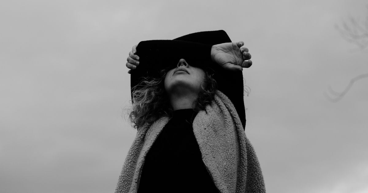 A black and white photo of a woman looking up to the sky. She is unsmiling and has raised her arms to cover the top of her face.