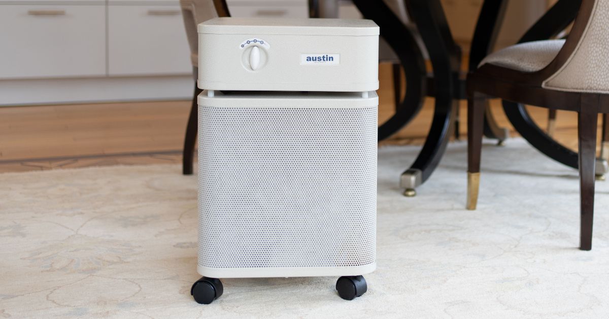 Sandstone standard size Austin Air purifier on a cream colored carpet in front of dining table.