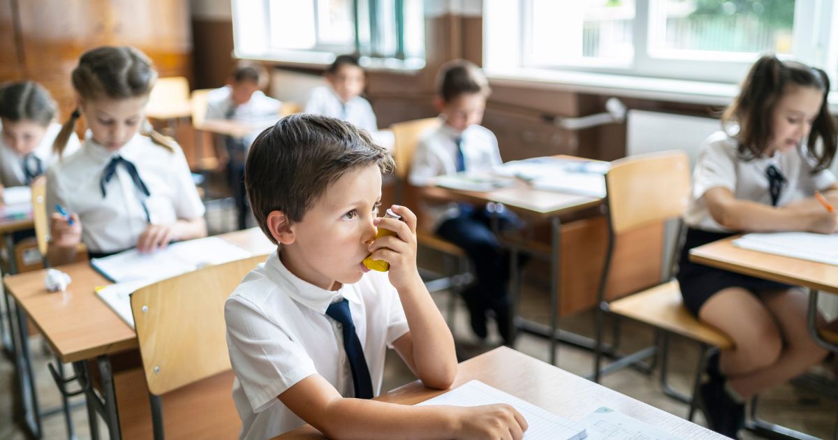 Young boy using his inhaler sitting at his desk in a classroom with other children also sitting at their desks.