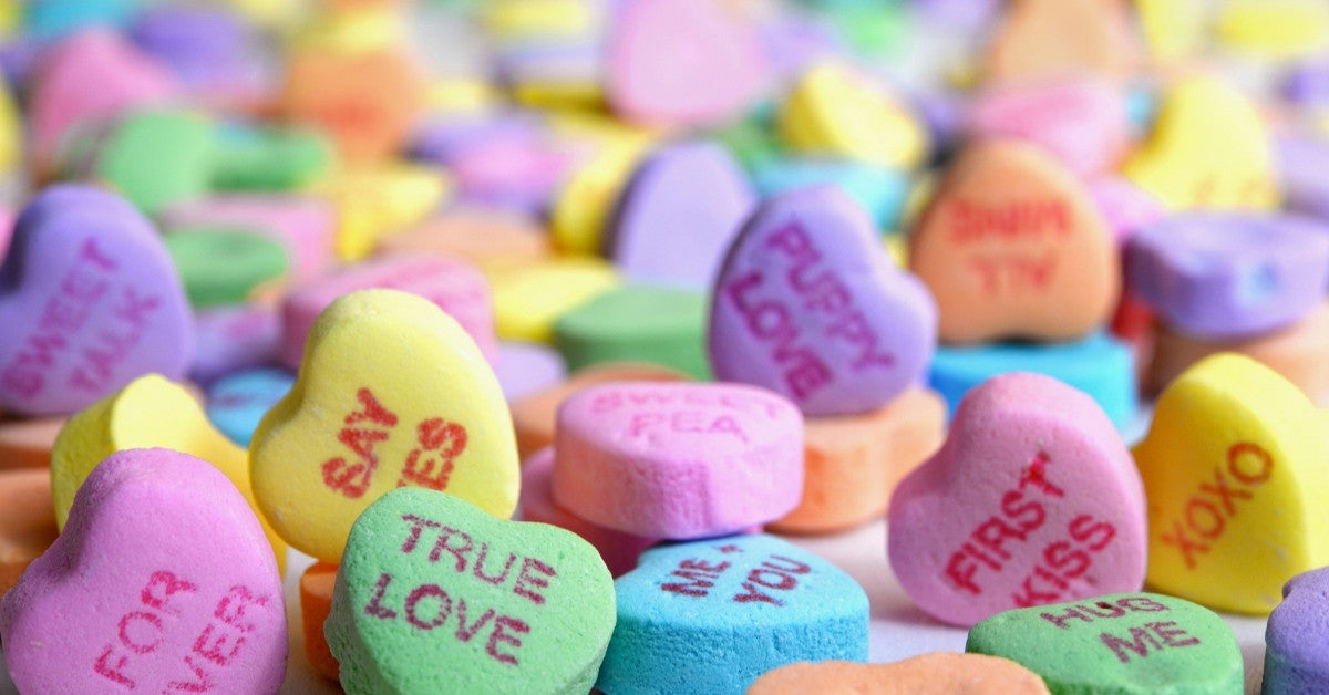 A close up photo of conversation hearts, the classic Valentine’s Day candy. They are small pastel hearts with words like, “true love,” “you plus me,” “hug me,” “puppy love,” and more. 
