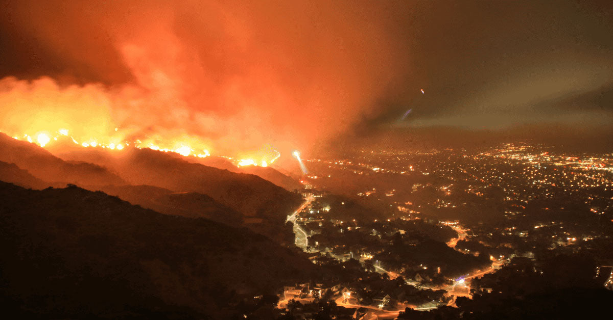 A landscape with a mountain on fire next to a city, with the flames moving from the left (in the mountains) toward the city. It is nighttime. 