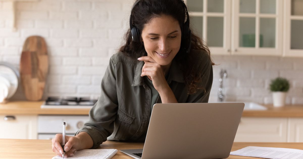 Woman wearing headphones is sitting in kitchen working on a laptop