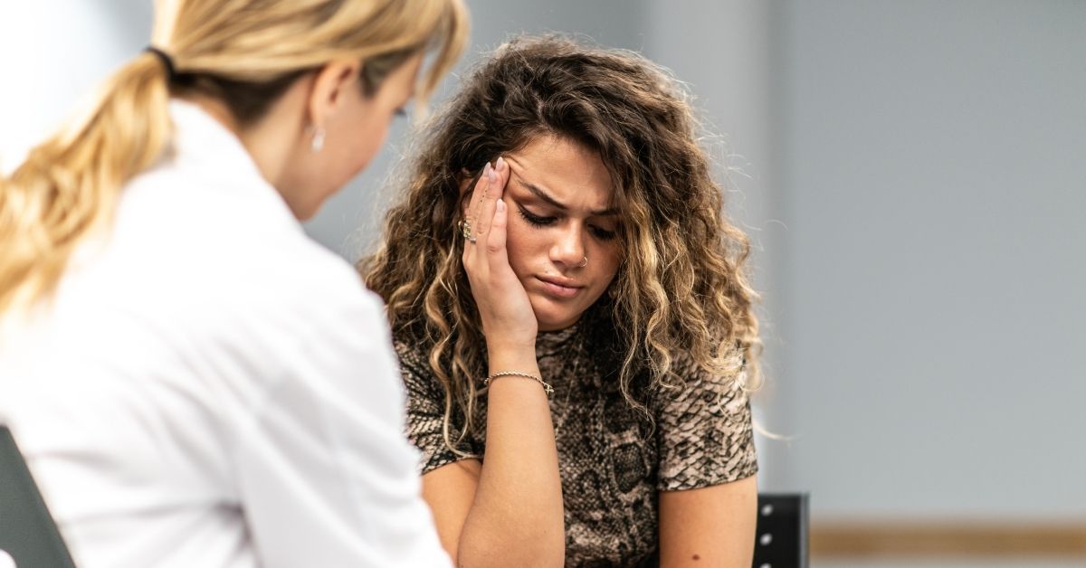 A photo of a woman with her hand held up to her face, she is looking unhappy and hurt. A doctor in a white coat is visible from behind, on the left.