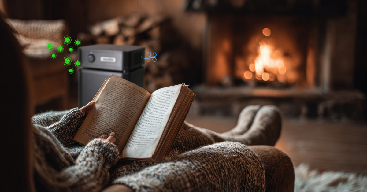 A photo showing a person sitting in a recliner, before a fire, reading a book. A black Austin Air Purifier is in the background with illustrations of green virus particles going into the unit and blue whiffs of fresh air coming out.