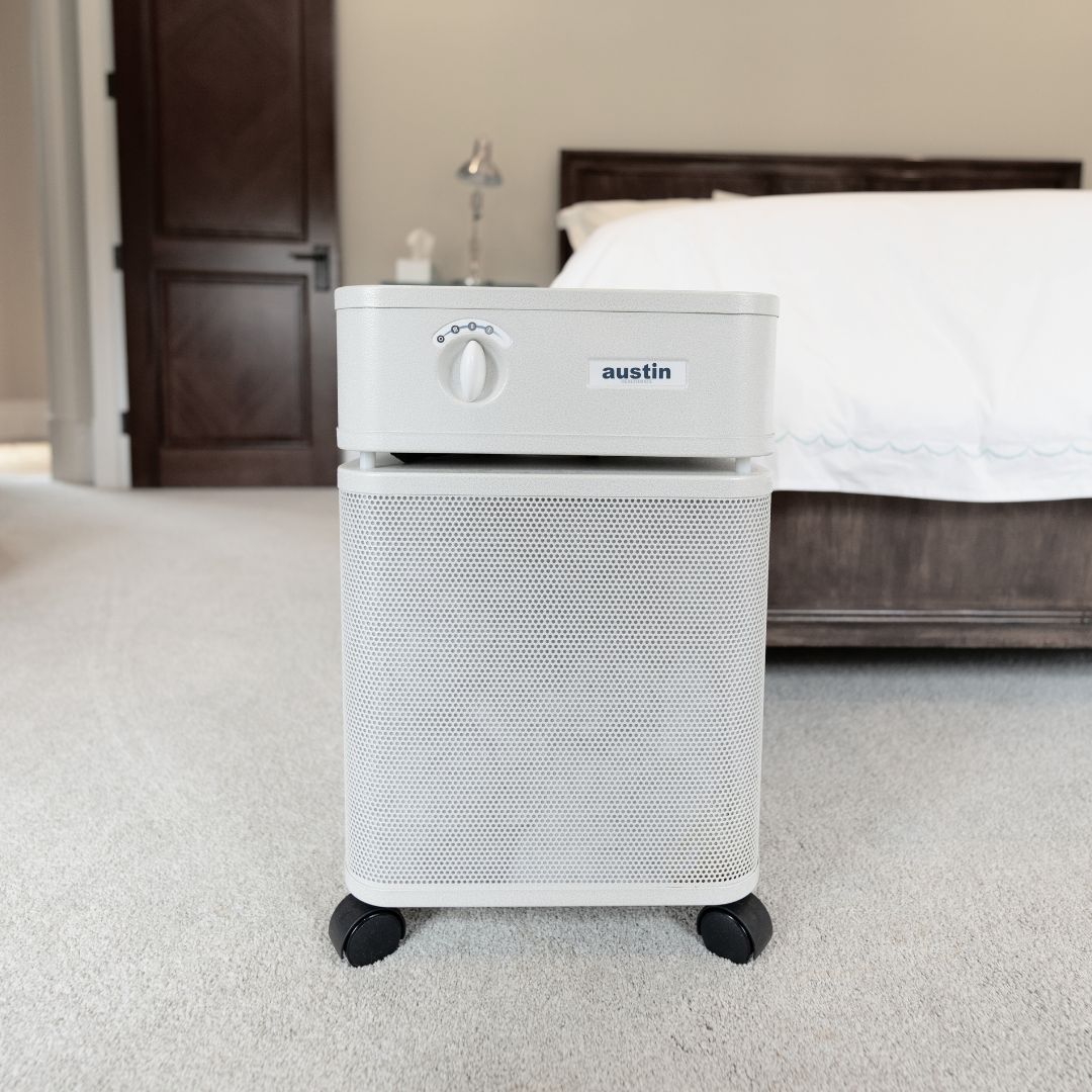White Austin Air purifier on a carpeted floor in a bedroom.