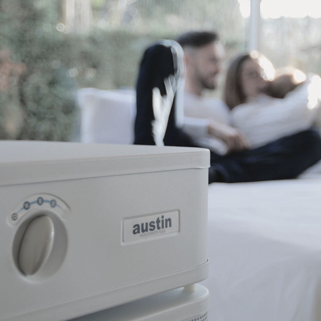 Close-up of a white Austin Air purifier, blurred background of mom, dad, and baby sitting