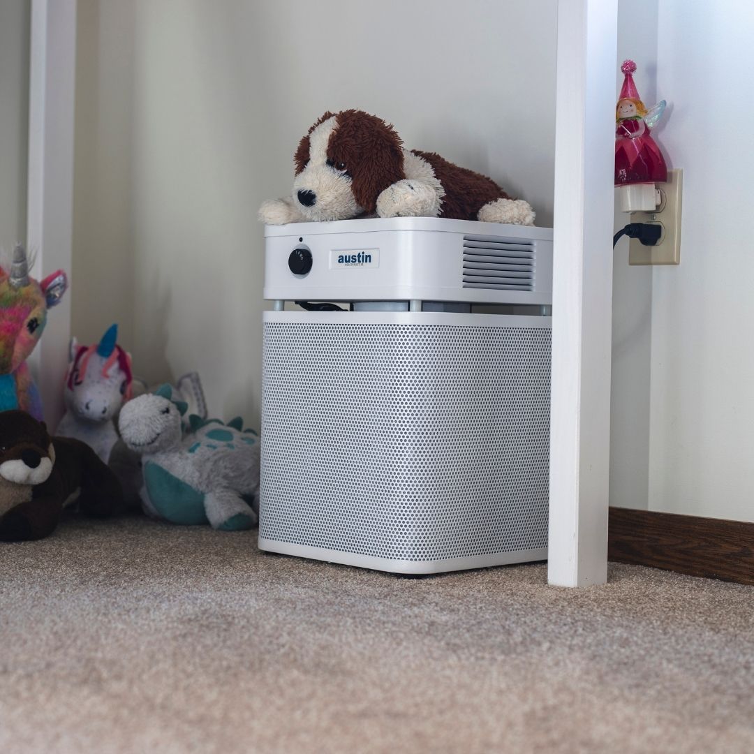 austin air healthmate plus junior air purifier with a plush dog toy on top in a room with stuffed animals.